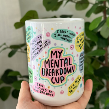 Close-up of a white ceramic affirmation mug with floral illustrations and bold text, held in hand with greenery background.