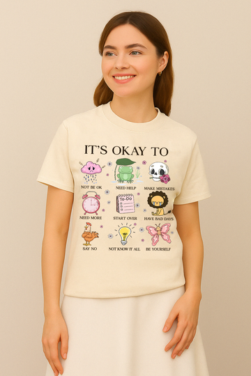 Ivory Comfort Colors t-shirt with “It’s Okay To Not Be Okay” design and colorful mental health icons. Worn by woman in skirt, smiling.