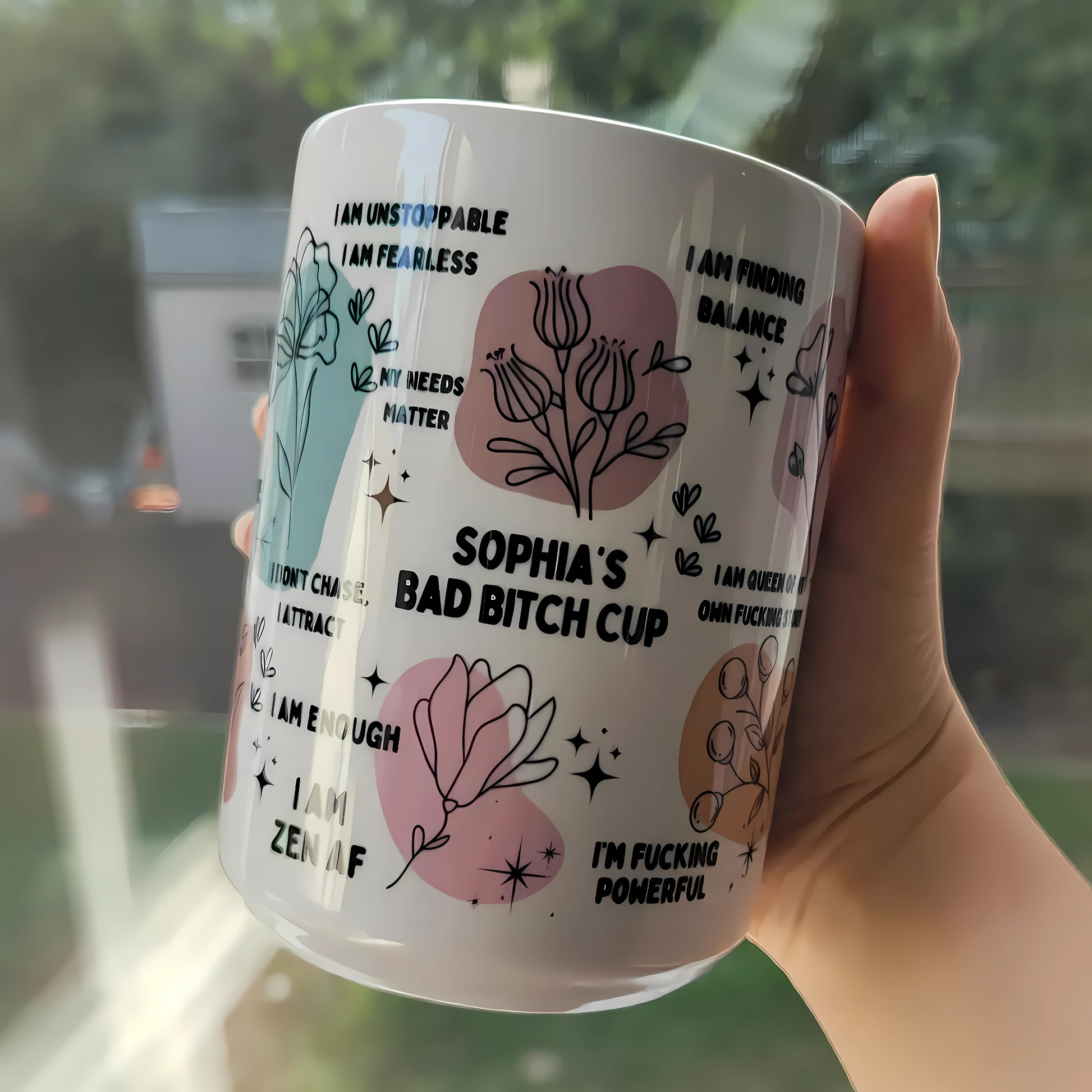 Close-up of a white ceramic affirmation mug with floral illustrations and bold text, held in hand with greenery background.