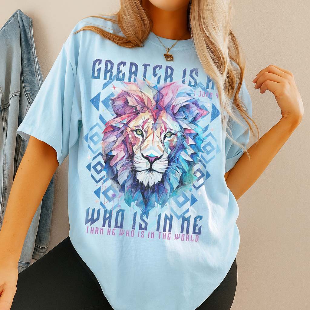 Model in chambray blue Comfort Colors t-shirt featuring vibrant geometric lion face and scripture “Greater Is He” – Christian faith tee.