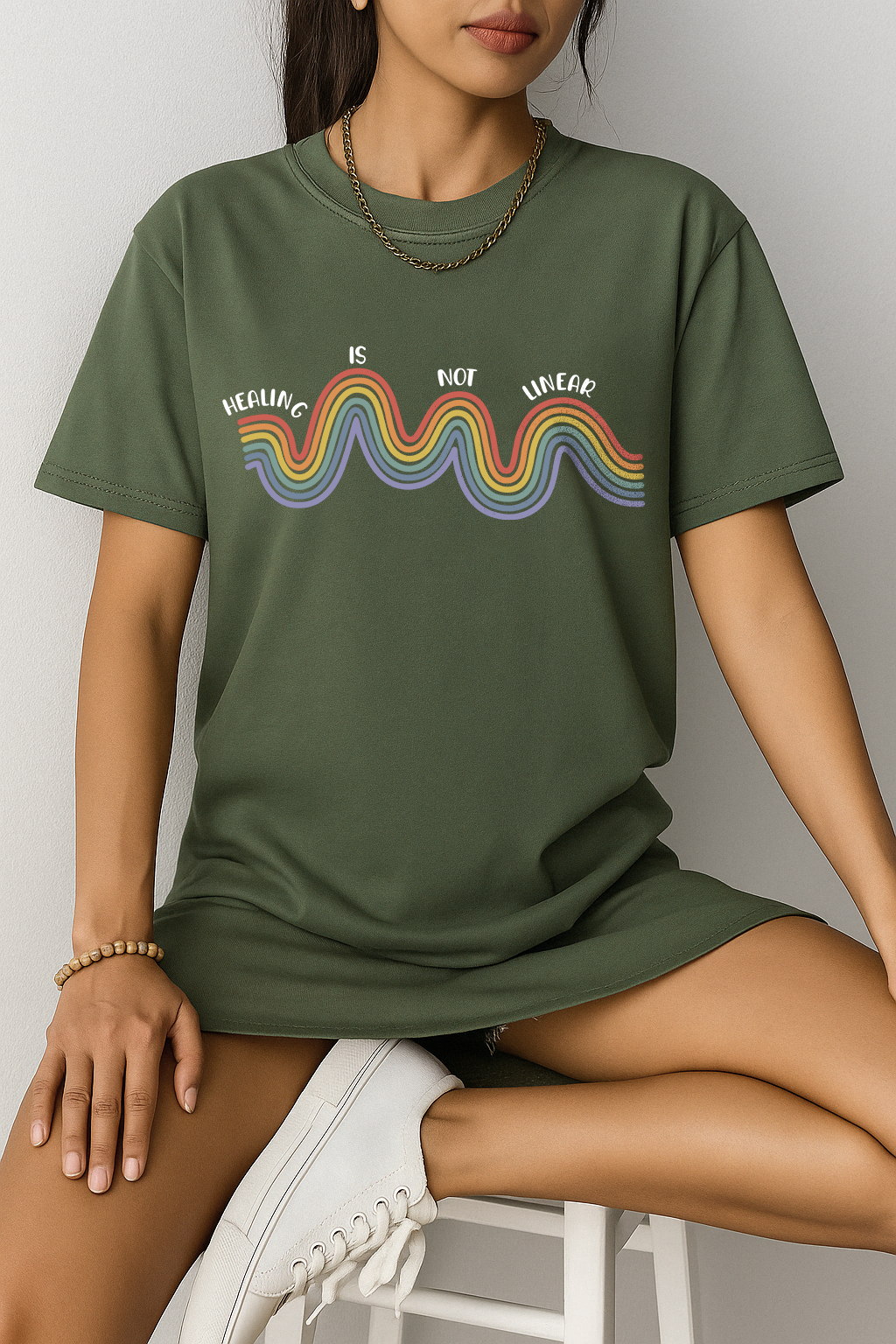Moss green Comfort Colors t-shirt with rainbow “Healing is not linear” quote. Worn by woman sitting on stool, casual mental health tee.