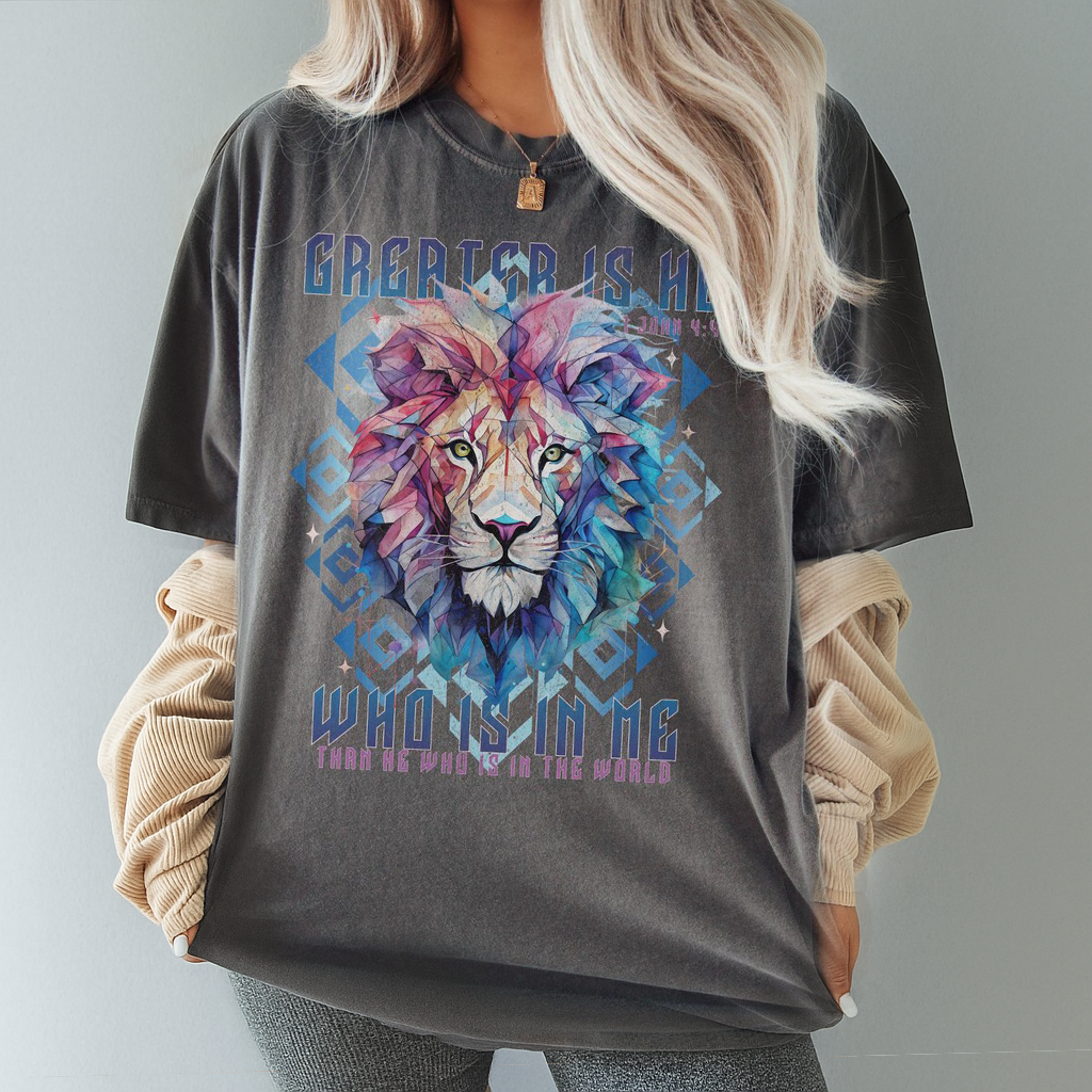 Relaxed-fit pepper color t-shirt styled casually with faith message “Greater Is He” and artistic lion face – Christian streetwear look.
