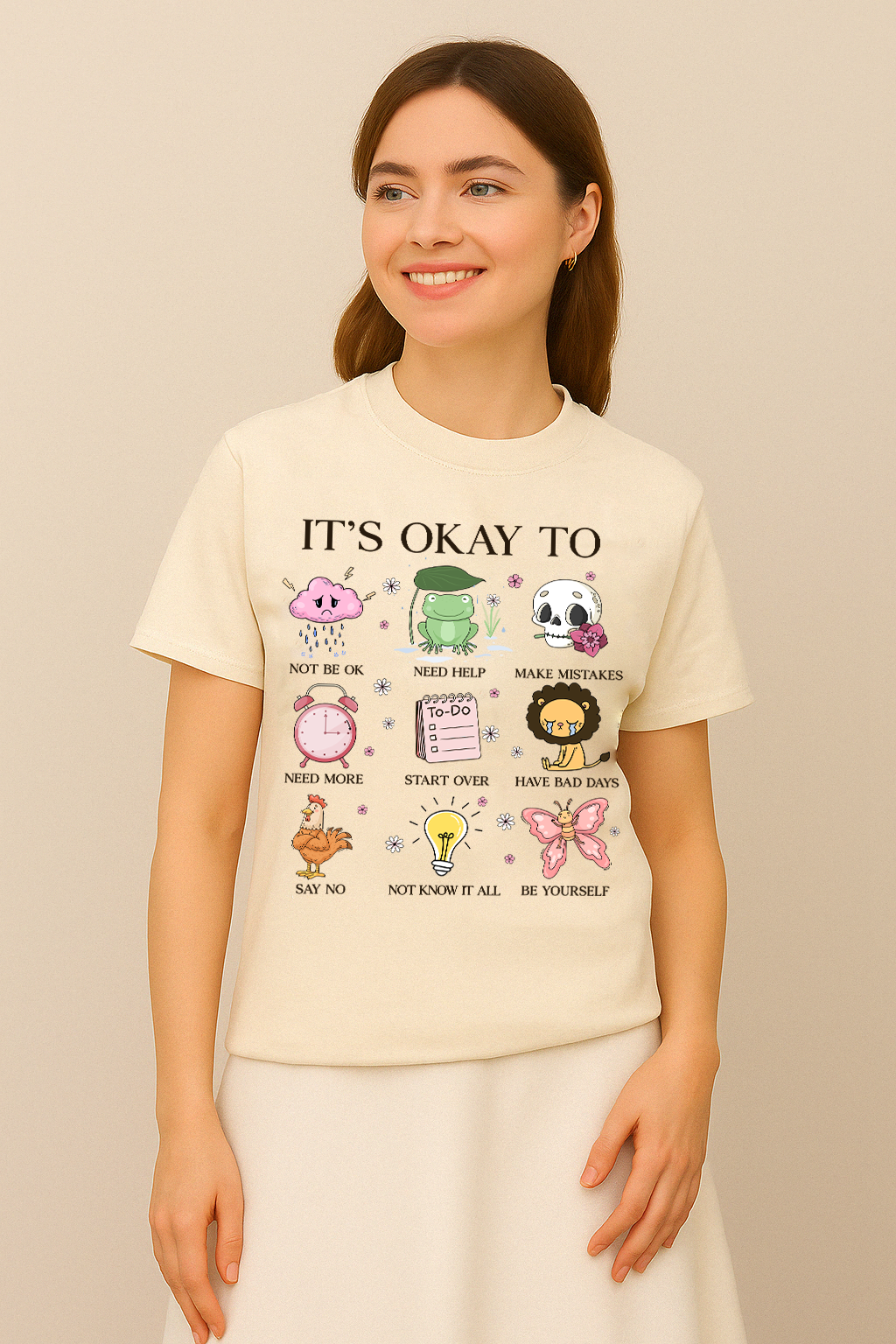 Ivory Comfort Colors t-shirt with “It’s Okay To Not Be Okay” design and colorful mental health icons. Worn by woman in skirt, smiling.