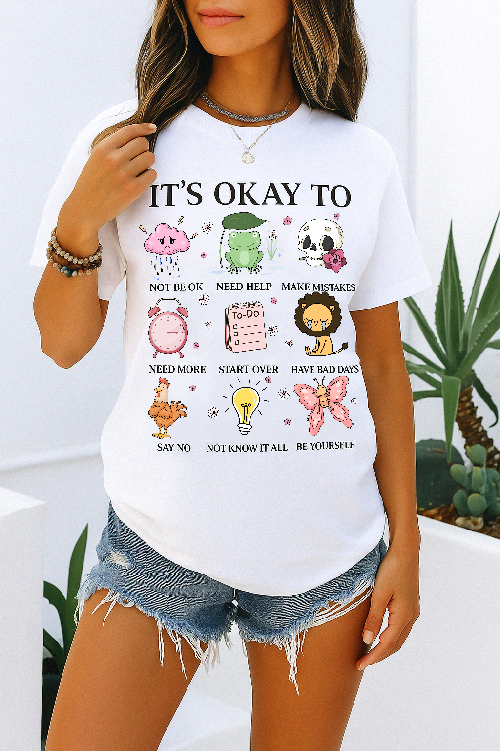 White Comfort Colors tee with “It’s Okay To Not Be Okay” quote and bright illustrated icons. Worn by woman in denim shorts, relaxed setting.